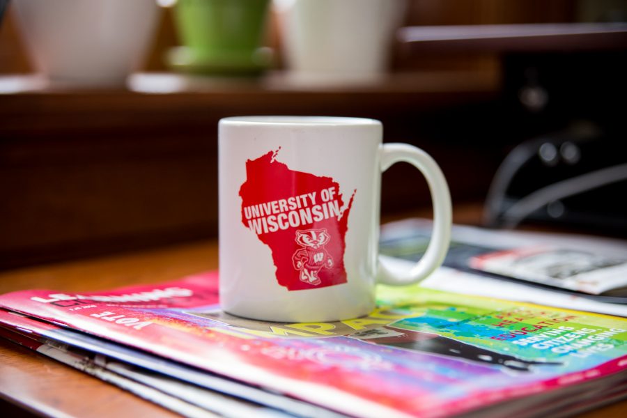 A white mug with an image of a red state of Wisconsin and Bucky the Badger on it atop of magazines.